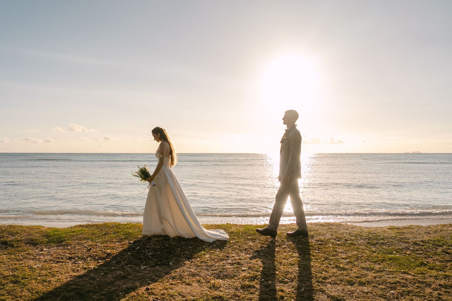 A couple in wedding attire stands silhouetted against a golden sunset on a scenic beach coastline.