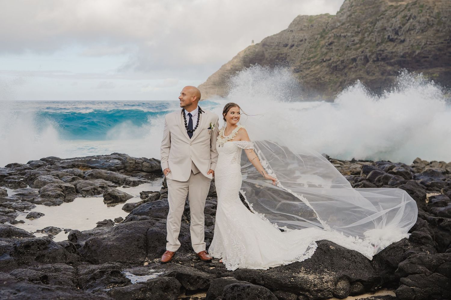 Bride and groom pose on rocky Hawaiian coastline with dramatic ocean waves and flowing wedding veil in the wind.