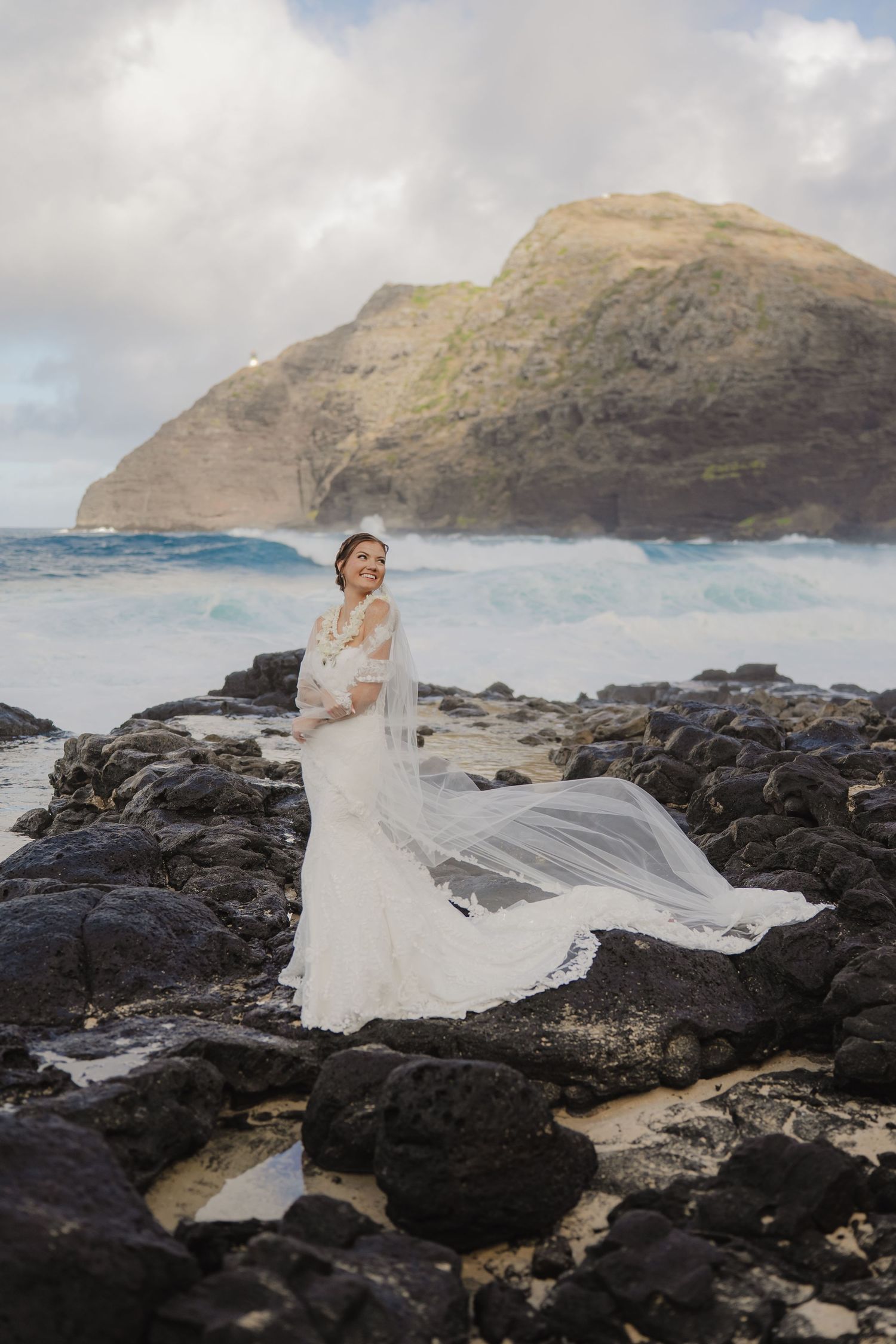 Bride in white lace gown with flowing veil on rocky Hawaii coastline with ocean waves and dramatic cliffs.