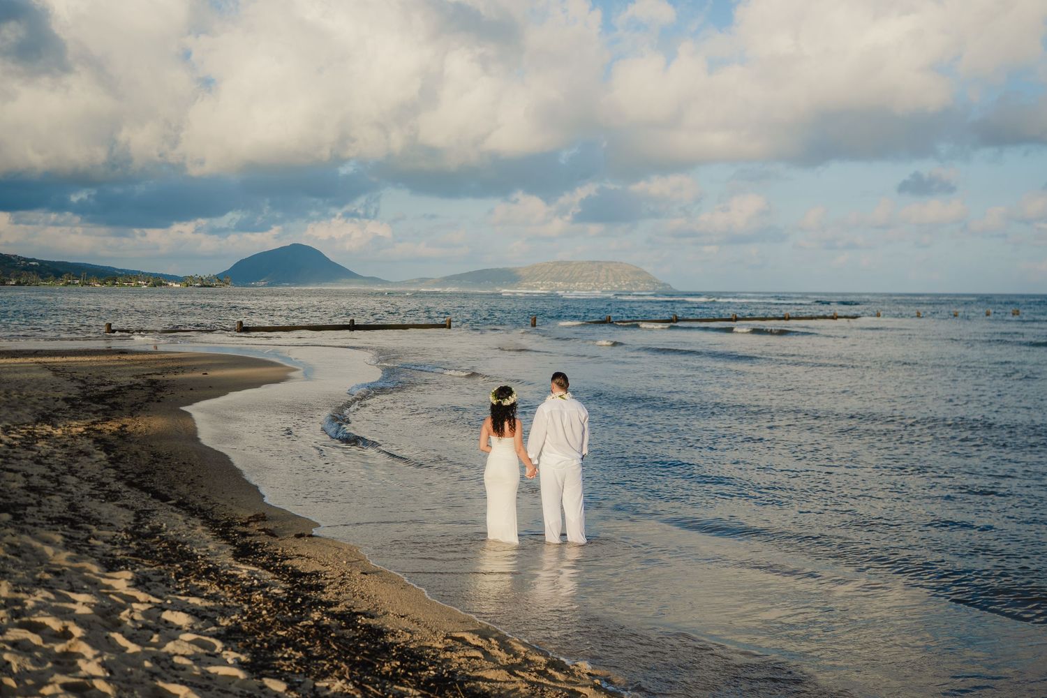 Couple in white attire walking hand-in-hand along a scenic beach with mountains and ocean in the background.