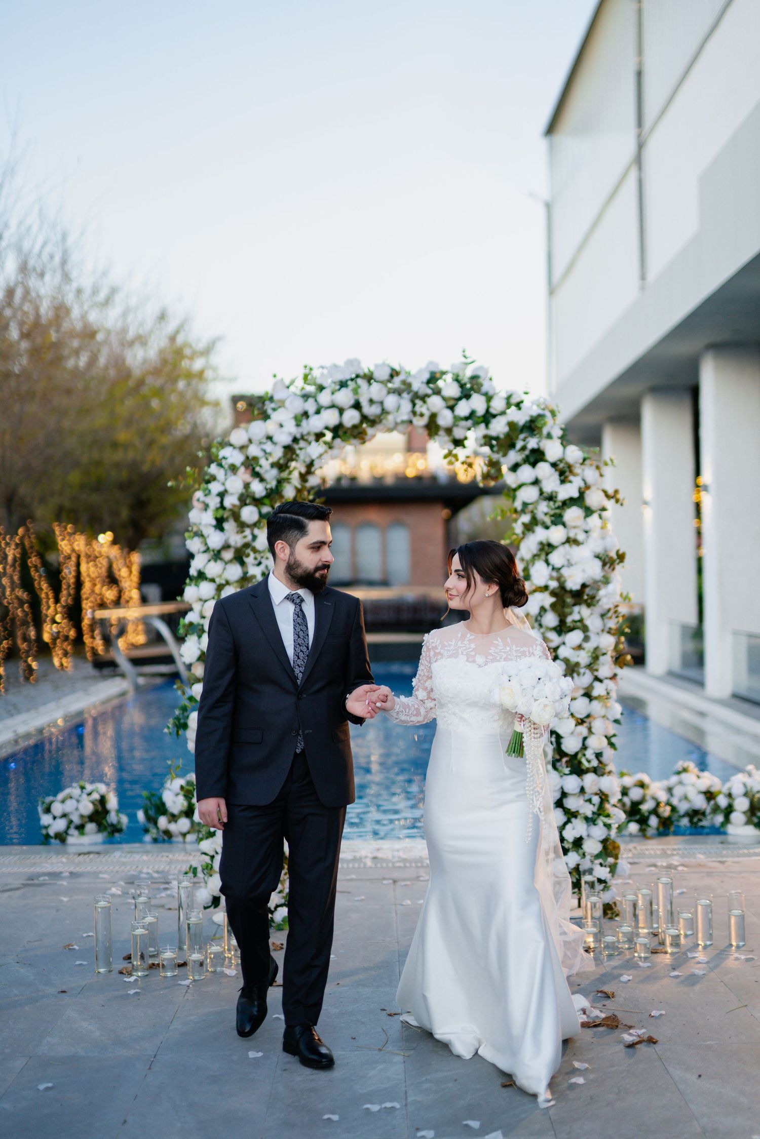 Elegant wedding couple walks together by a pool under a beautiful floral arch decorated with white flowers at dusk.