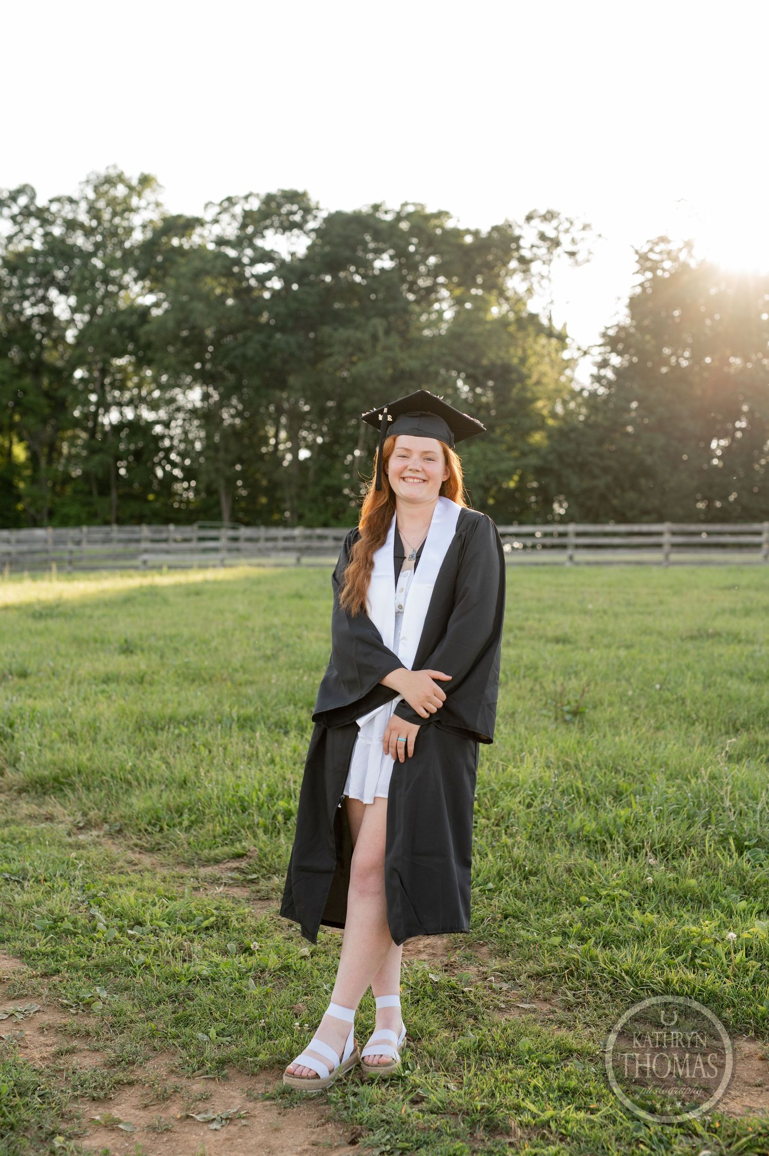 Graduate poses in academic regalia on farm at sunset.