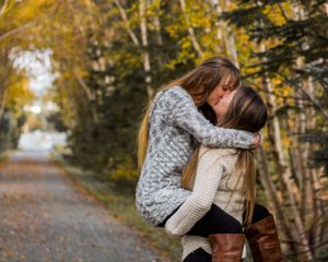 A couple in cozy sweaters embrace on an autumn path surrounded by golden trees and fallen leaves.
