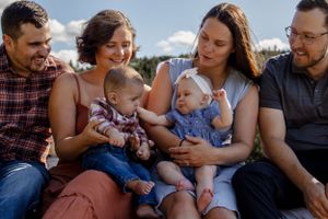 A group sits together outdoors on a sunny day with two small babies wearing blue and plaid outfits.