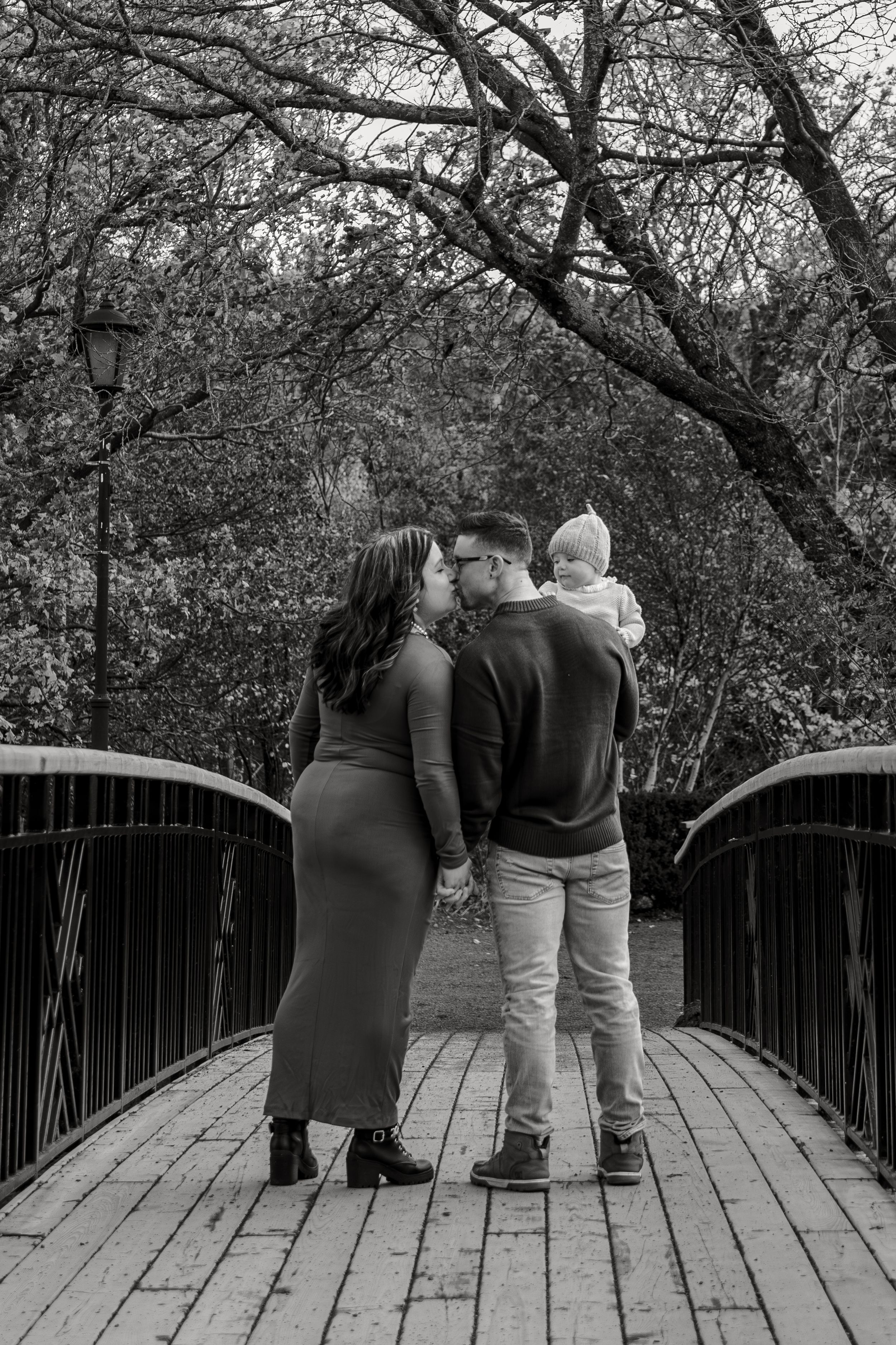 A couple stands together on a wooden bridge in a park setting, captured in black and white photography.