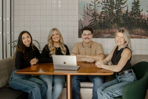 A group sits together at a cafe table with a laptop, dressed casually in jeans against a wall with forest artwork.