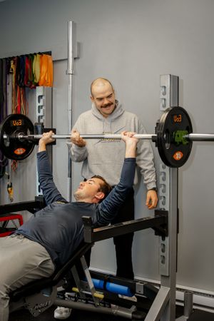A weightlifter performs bench press exercises with a spotter standing nearby in a well-equipped gym.