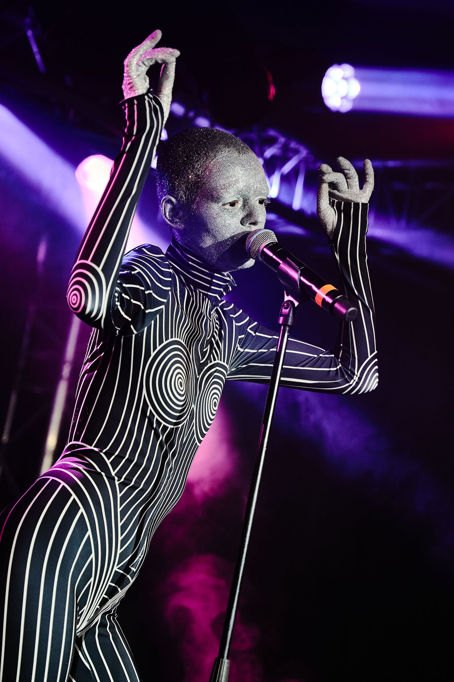 Performer wearing striking black and white spiral bodysuit sings into microphone on stage with dramatic purple lighting.