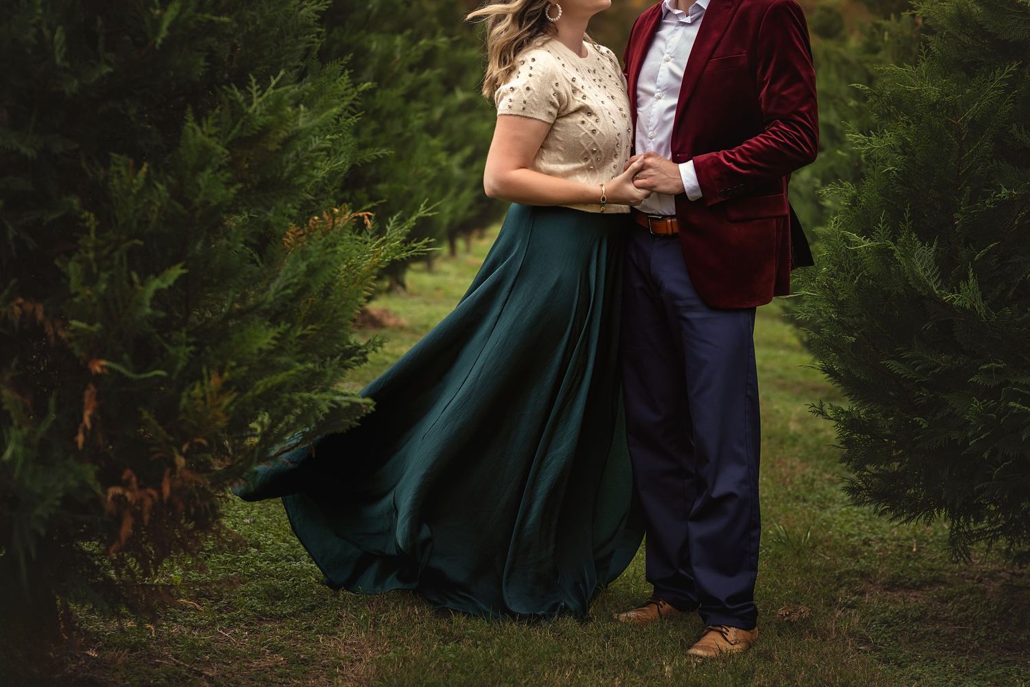 A couple in vintage-inspired formal wear embraces in a Christmas tree farm, her emerald green skirt flowing against his burgundy jacket.