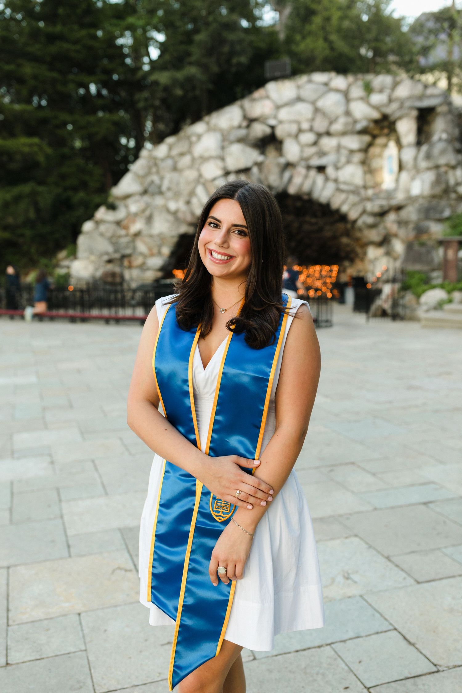 A graduation photo showing someone in a blue and gold academic stole and white dress standing in front of a stone archway.