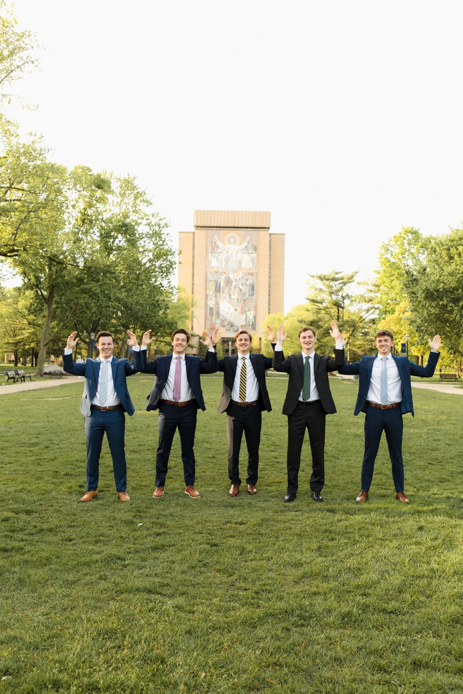 Five graduates in dark suits standing on grass lawn in front of university building.