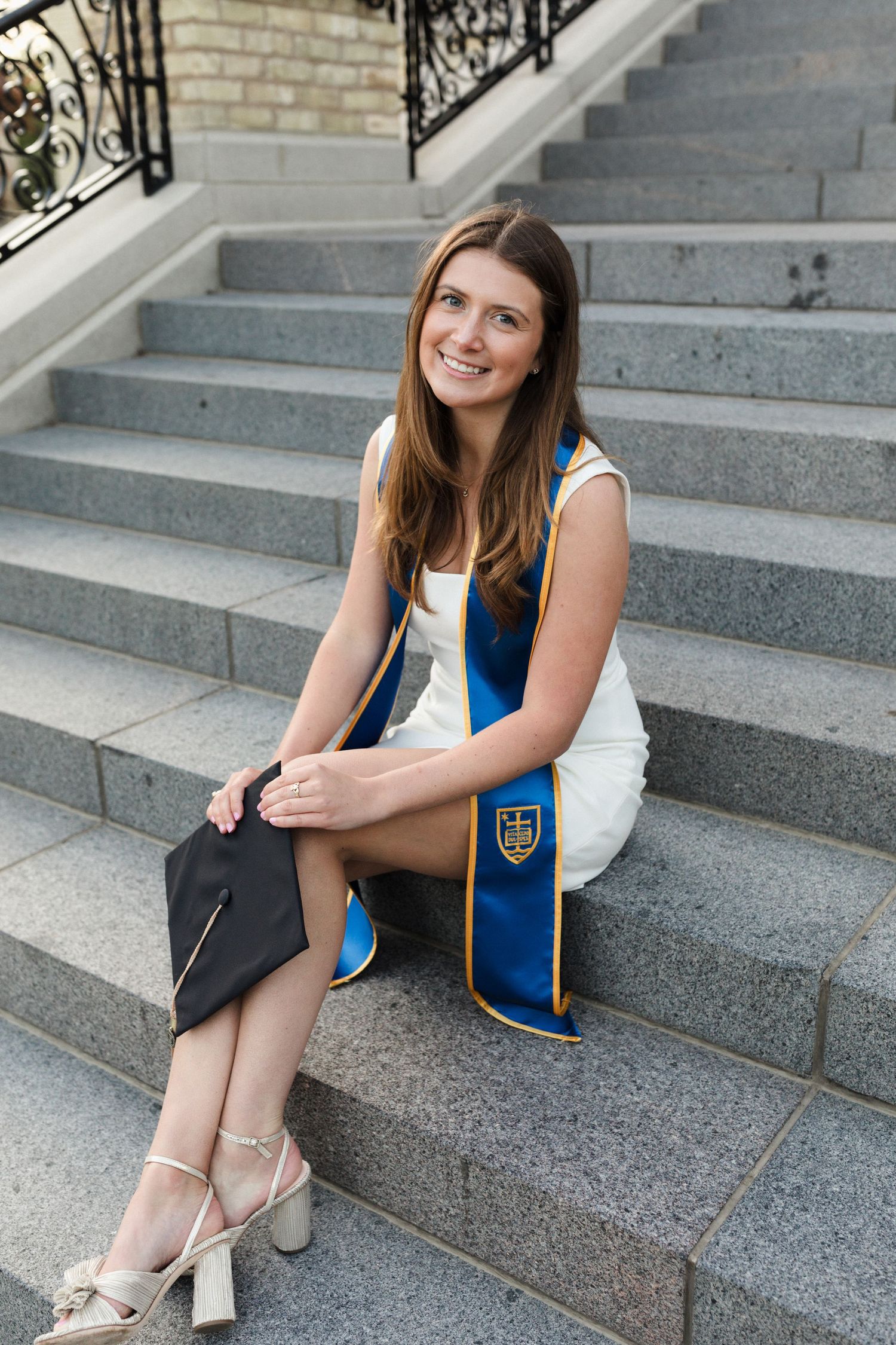 Graduate in white dress and blue stole sitting on grand stone staircase.
