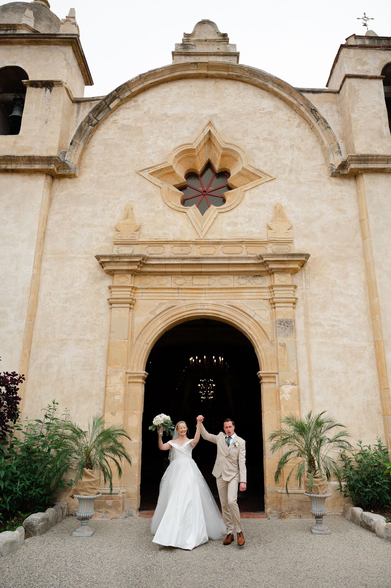 Two wedding attendees pose against a rustic wooden fence with climbing ivy and brick patio in a garden setting.