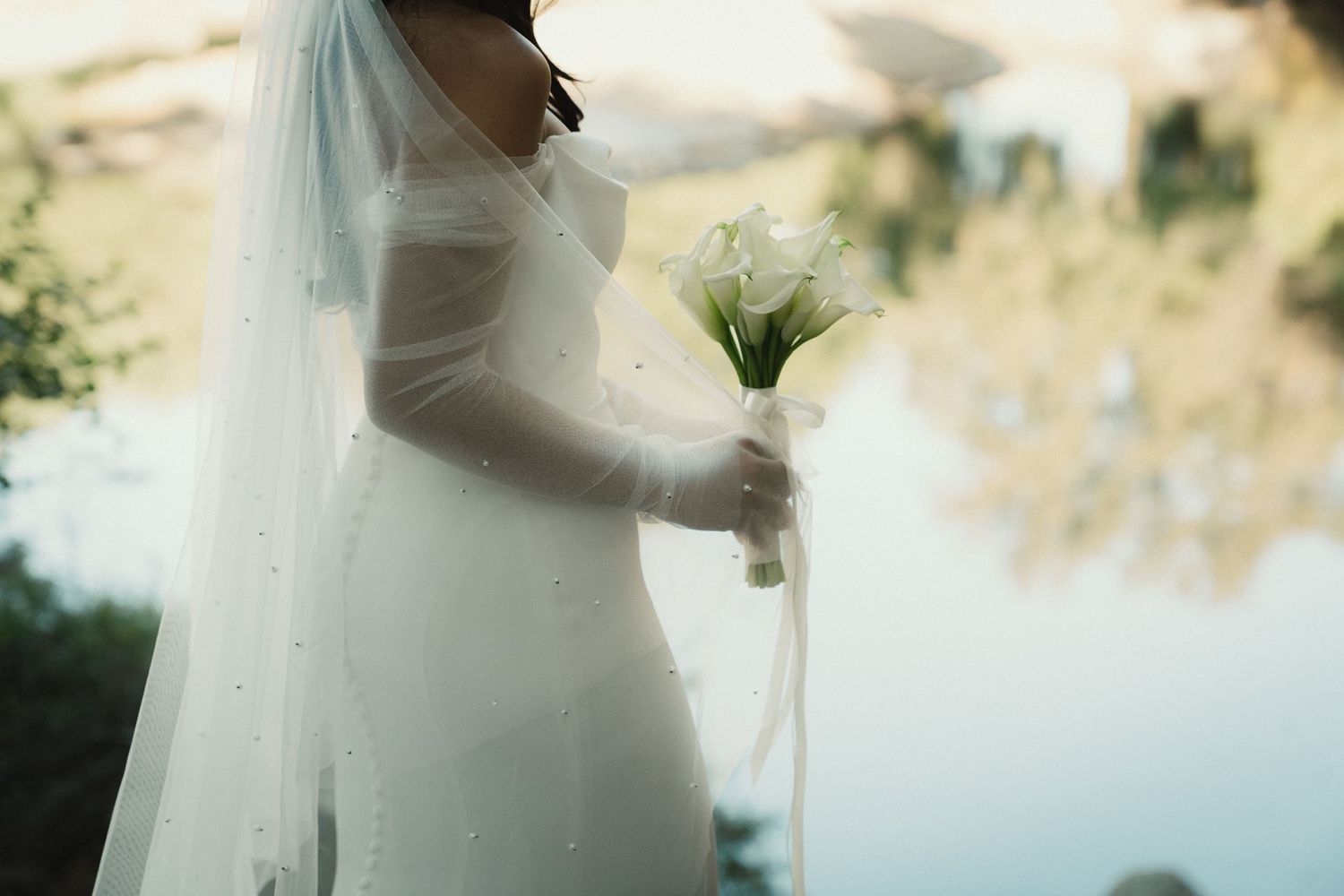 Dreamy close-up of a bride in a flowing white dress holding a white bouquet against soft sunlit background.