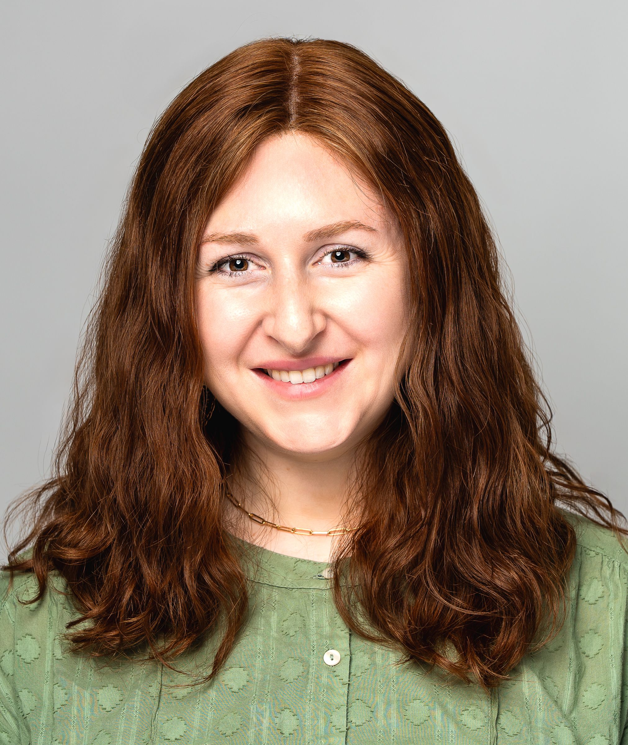 Headshot portrait of a woman with long wavy brown burnt hair wearing a green top and delicate gold necklace, smiling warmly at camera.