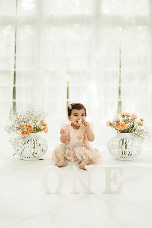 A baby sits among delicate floral arrangements and glass vases on a white surface.