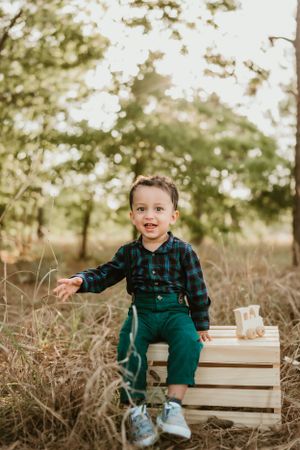 Child in plaid shirt and overalls sits on wooden crate during outdoor autumn photo session.