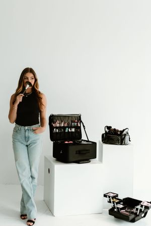 Woman in casual denim poses with professional makeup cases in white studio setting.