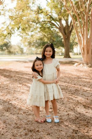 Two children wearing matching white dresses stand together under autumn trees in a park setting.