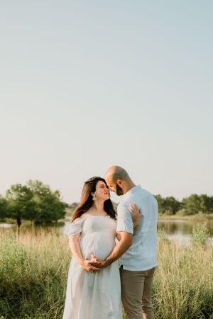 A couple embraces in a sunlit field wearing light colored clothing during a maternity photoshoot.