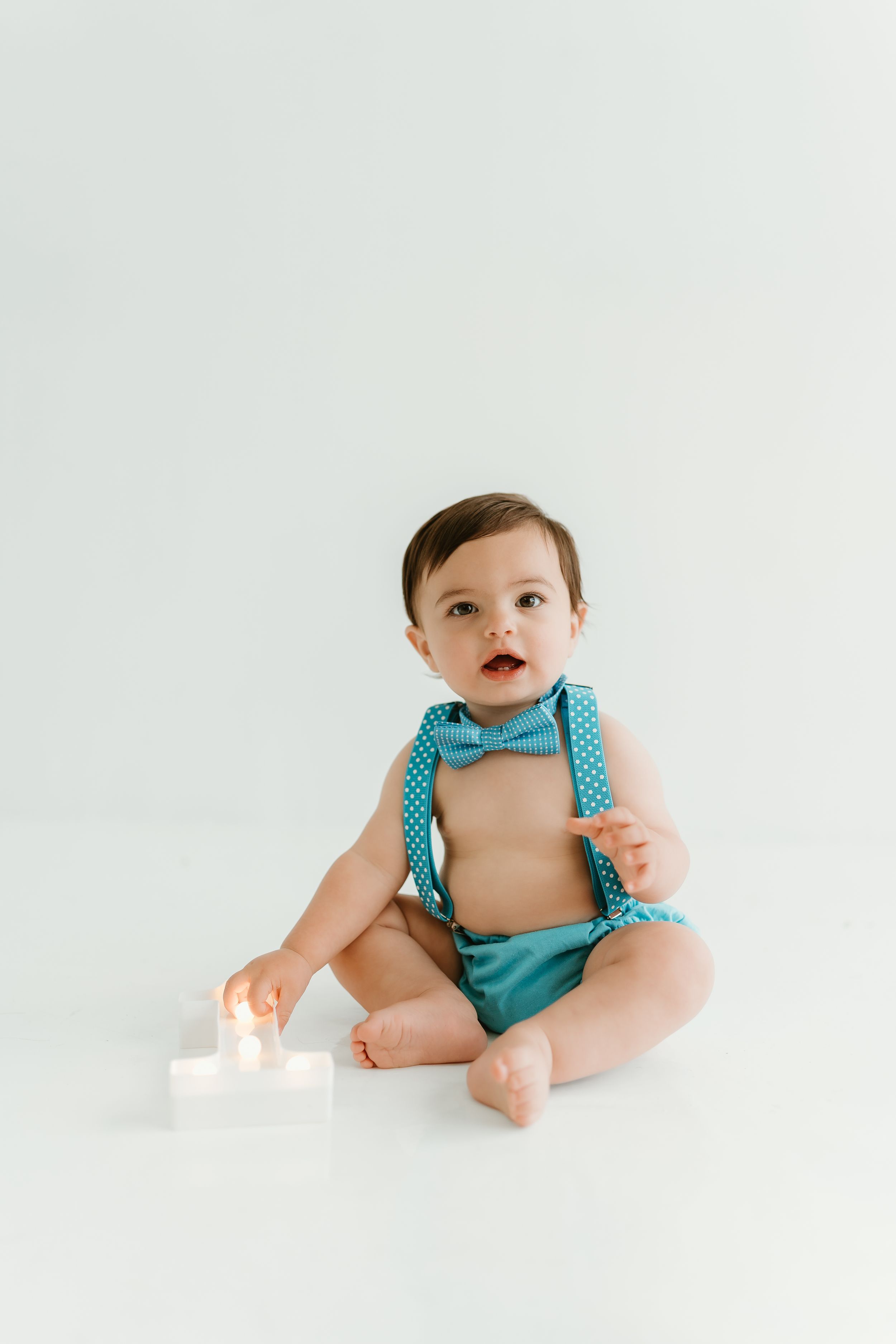 Baby wearing turquoise romper sits and smiles during minimalist studio photoshoot.