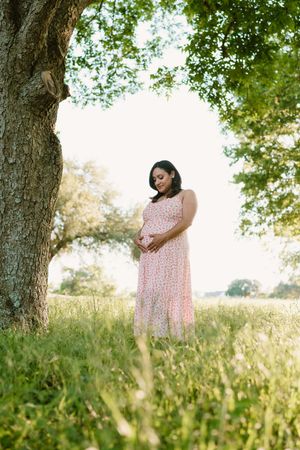 Woman in pink floral dress poses gracefully in a sunlit meadow with trees in the background.