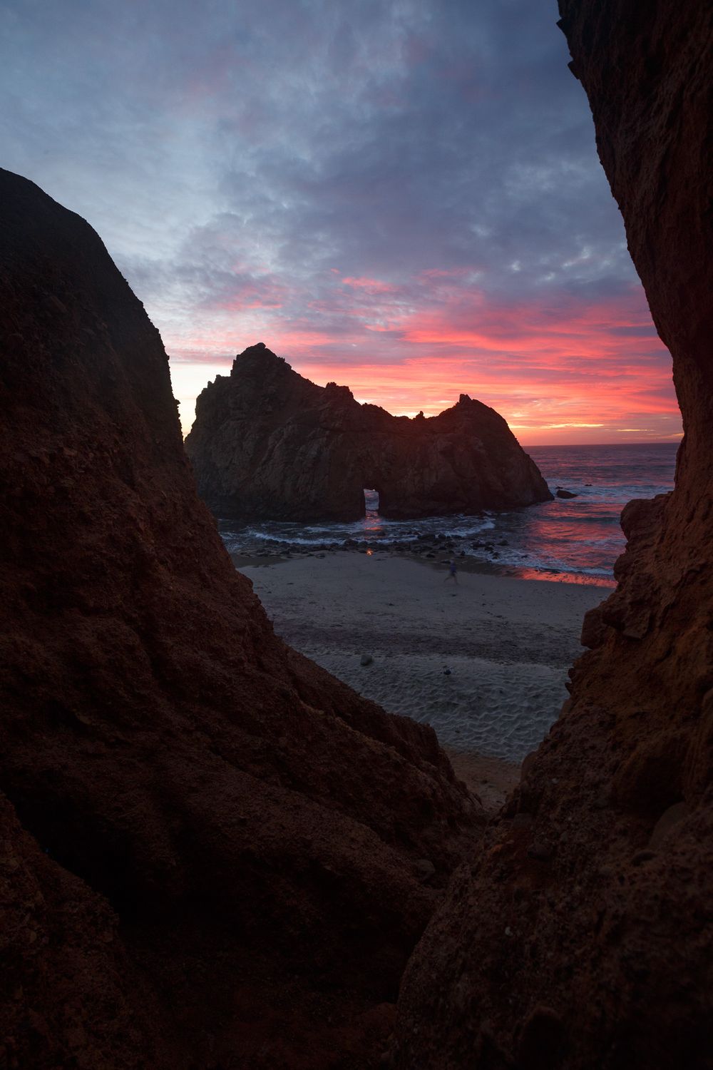 A lighthouse stands between rocky cliffs on a beach at sunset with vibrant pink and purple clouds in the sky.