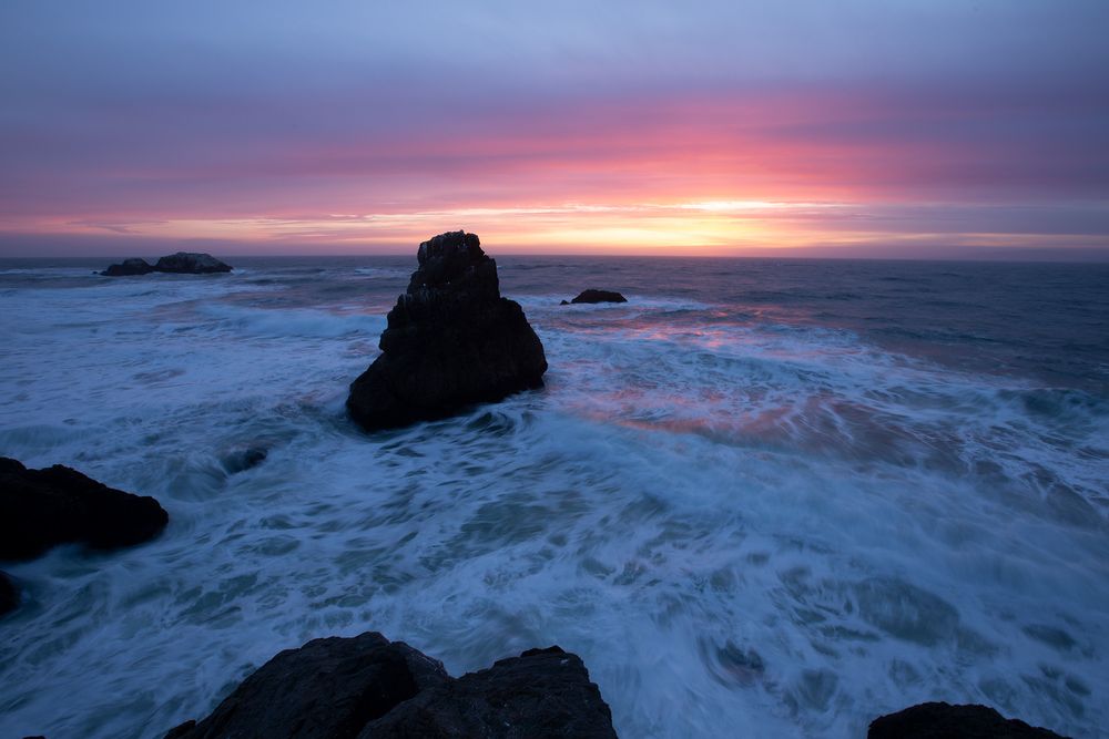 Dramatic sunset illuminates crashing waves against dark coastal rocks as twilight settles over the rugged ocean shoreline.