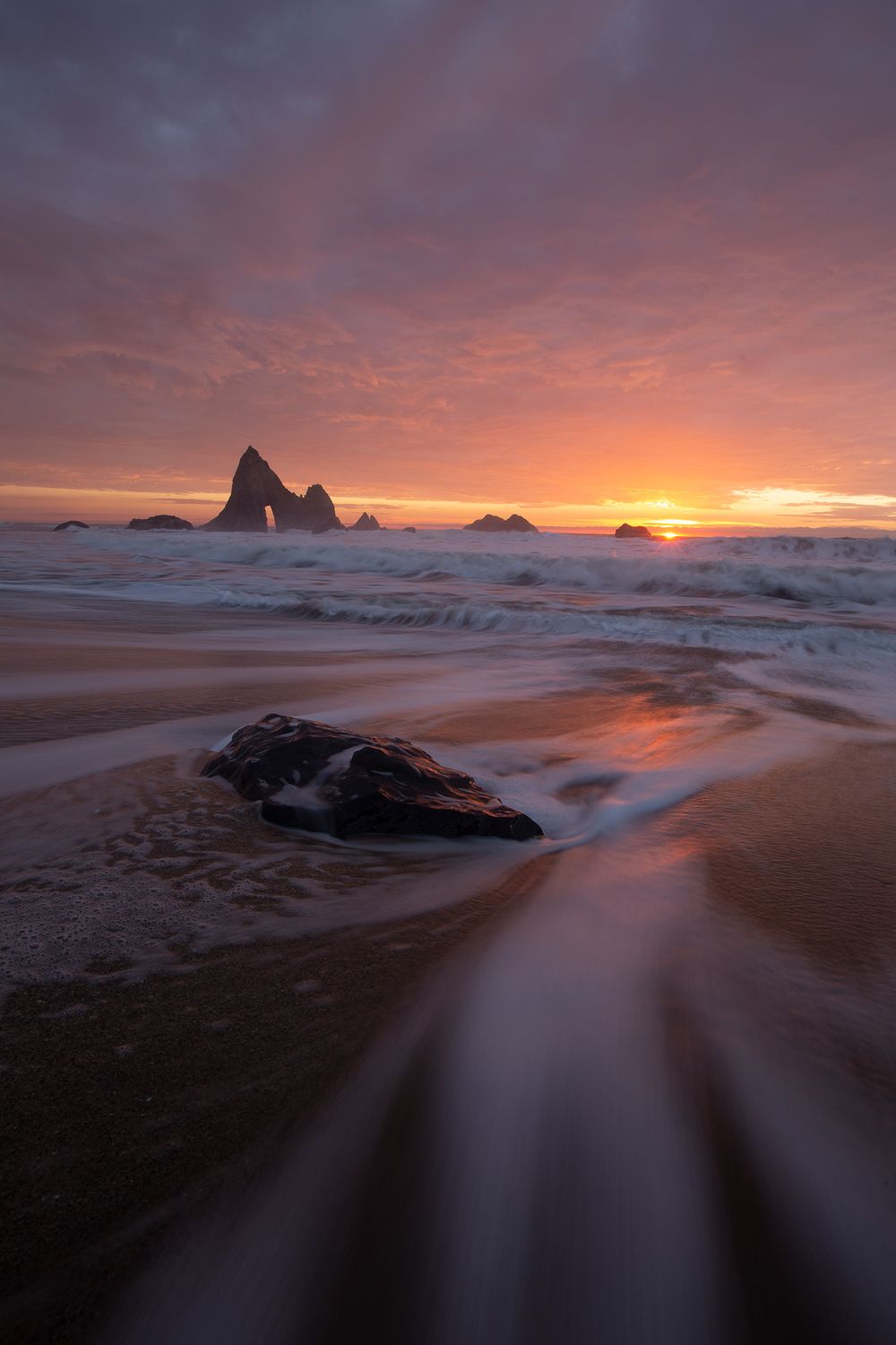 Dramatic coastal sunset illuminates rugged rock formations and flowing water on a beach with vibrant orange sky.
