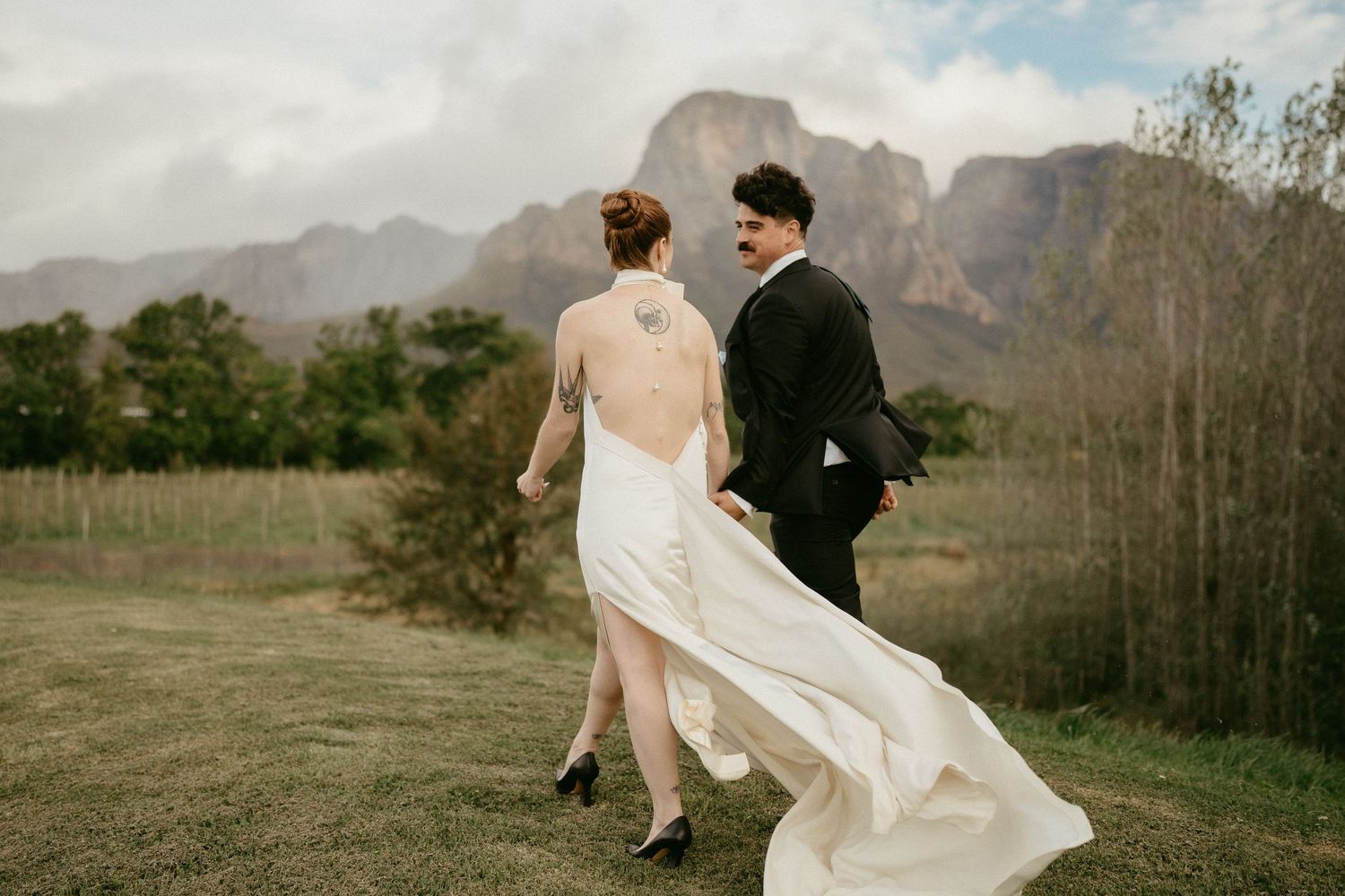 A couple in wedding attire silhouetted against dramatic mountain peaks and a cloudy sky in a scenic outdoor landscape.