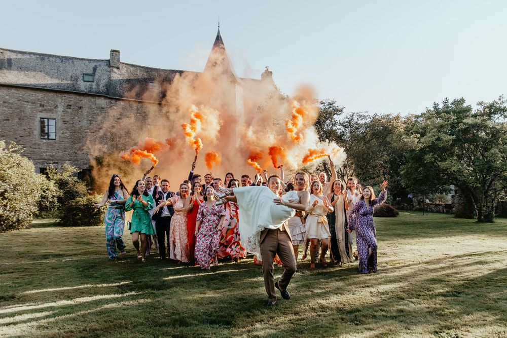 Wedding group photo with orange smoke bombs in front of a historic stone building.