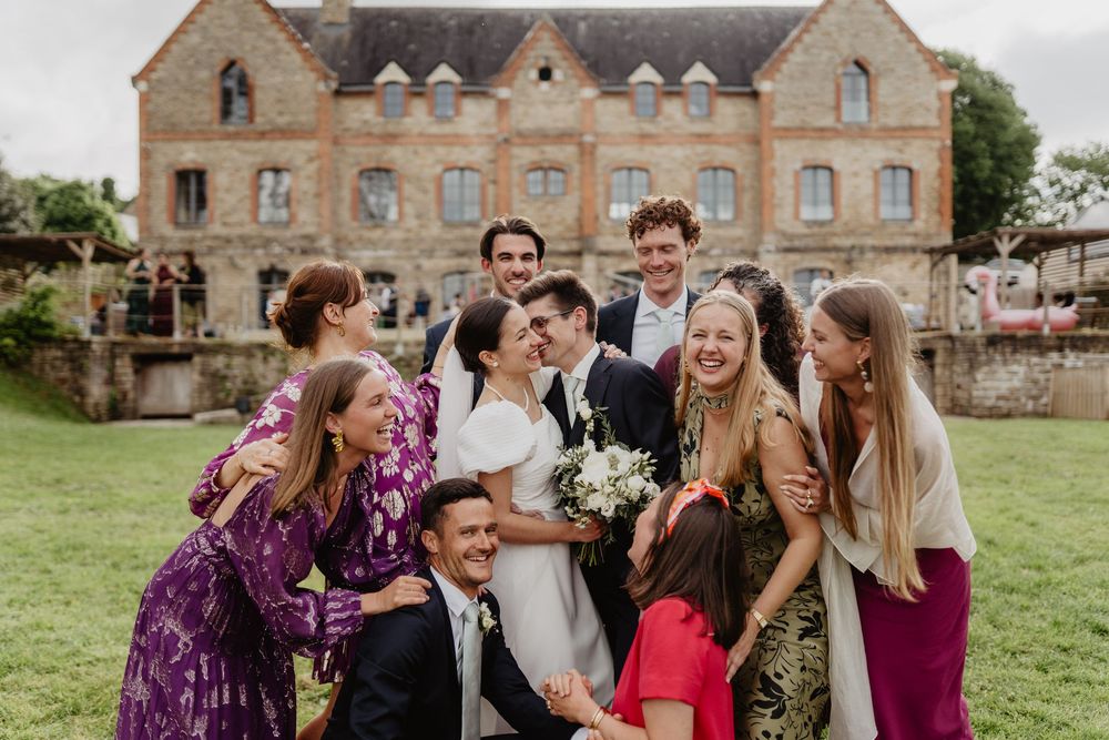 Bride and groom surrounded by joyful wedding guests laughing and embracing outside a historic stone mansion.