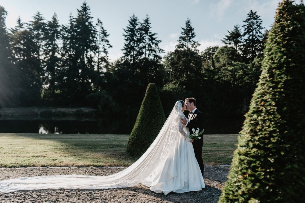 Bride and groom kissing in a lush garden with tall hedges and trees, bride wearing a long veil trailing on grass.