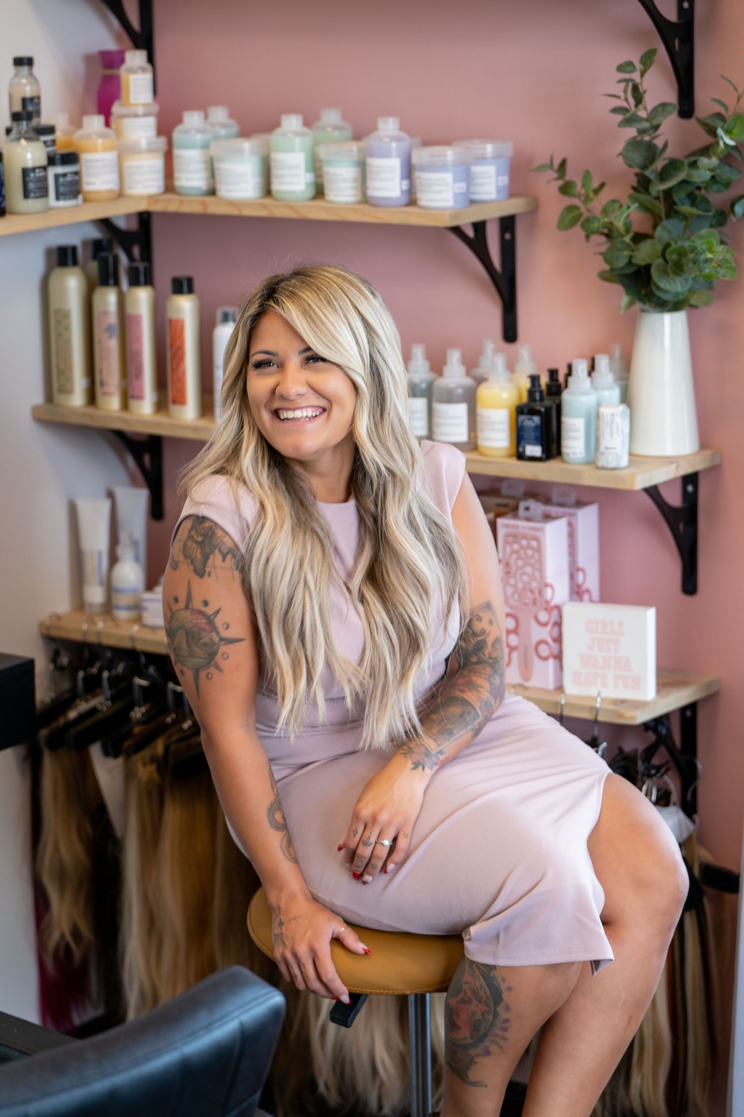 Hair salon interior with pink walls, wooden shelving displaying beauty products, and a styling chair in view.