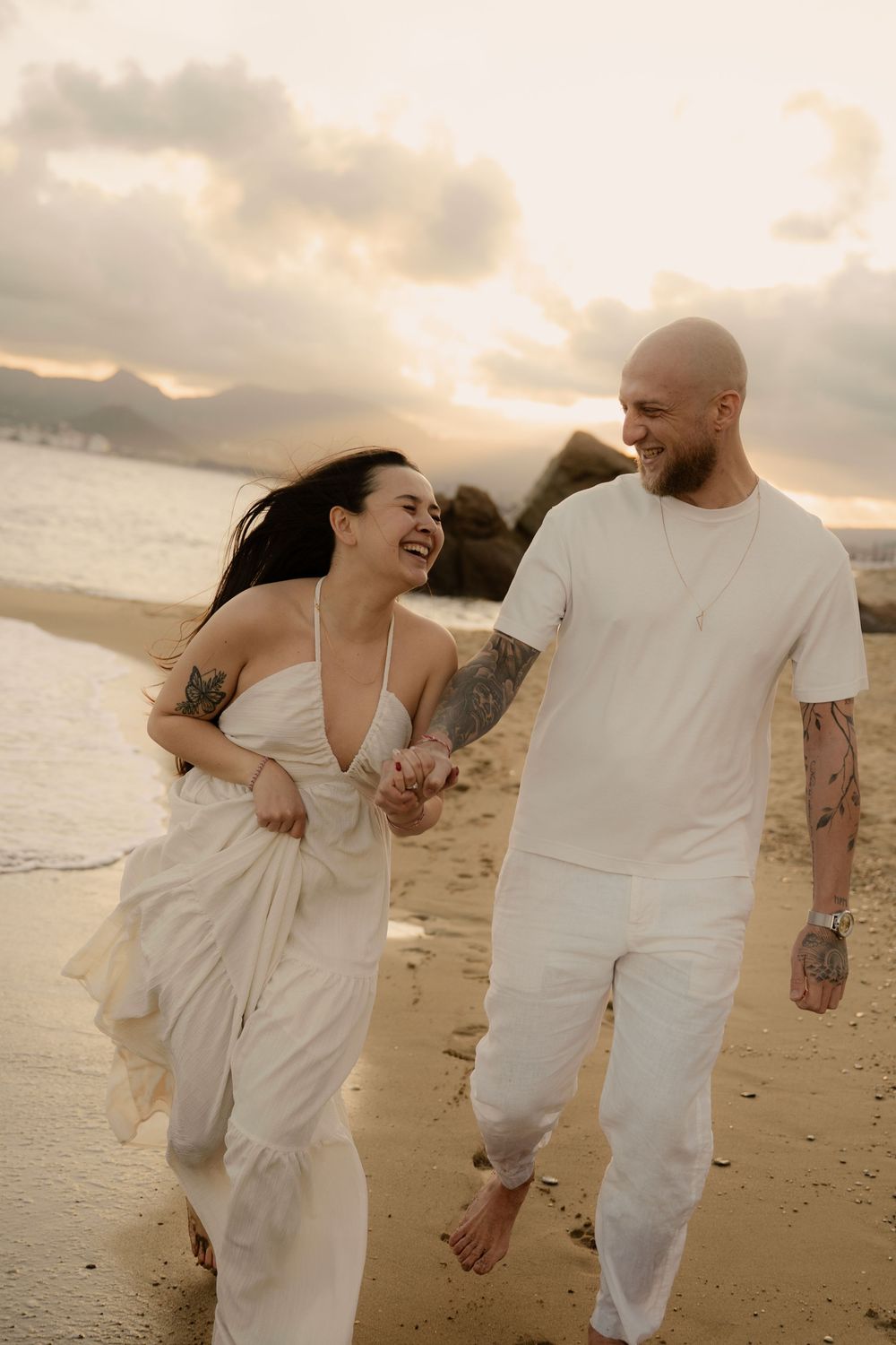 Un couple vêtu de blanc courent en riant main dans la main sur une plage au coucher du soleil.