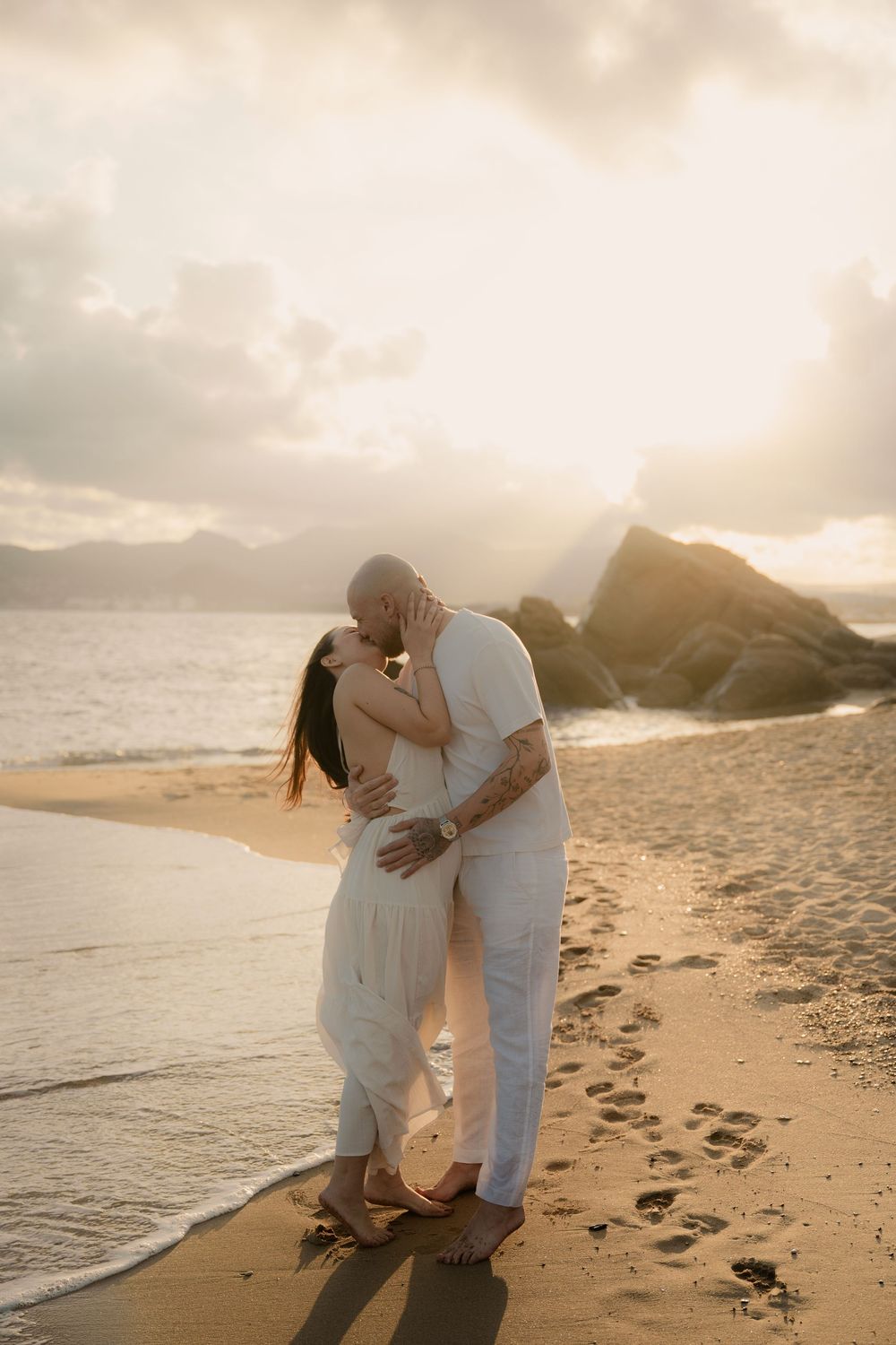 Un couple romantique en tenues blanches s'enlace sur une plage de sable au coucher du soleil, avec les vagues et les rochers en arrière-plan