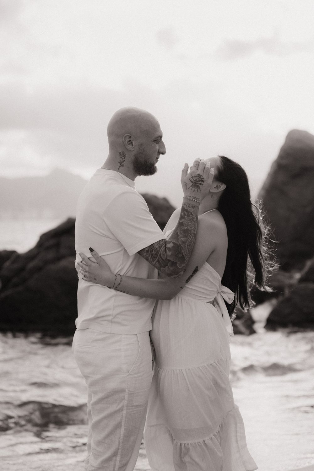 Une photo romantique en noir et blanc sur une plage, montrant un couple enlacé, vêtu de blanc, devant un décor côtier rocheux.
