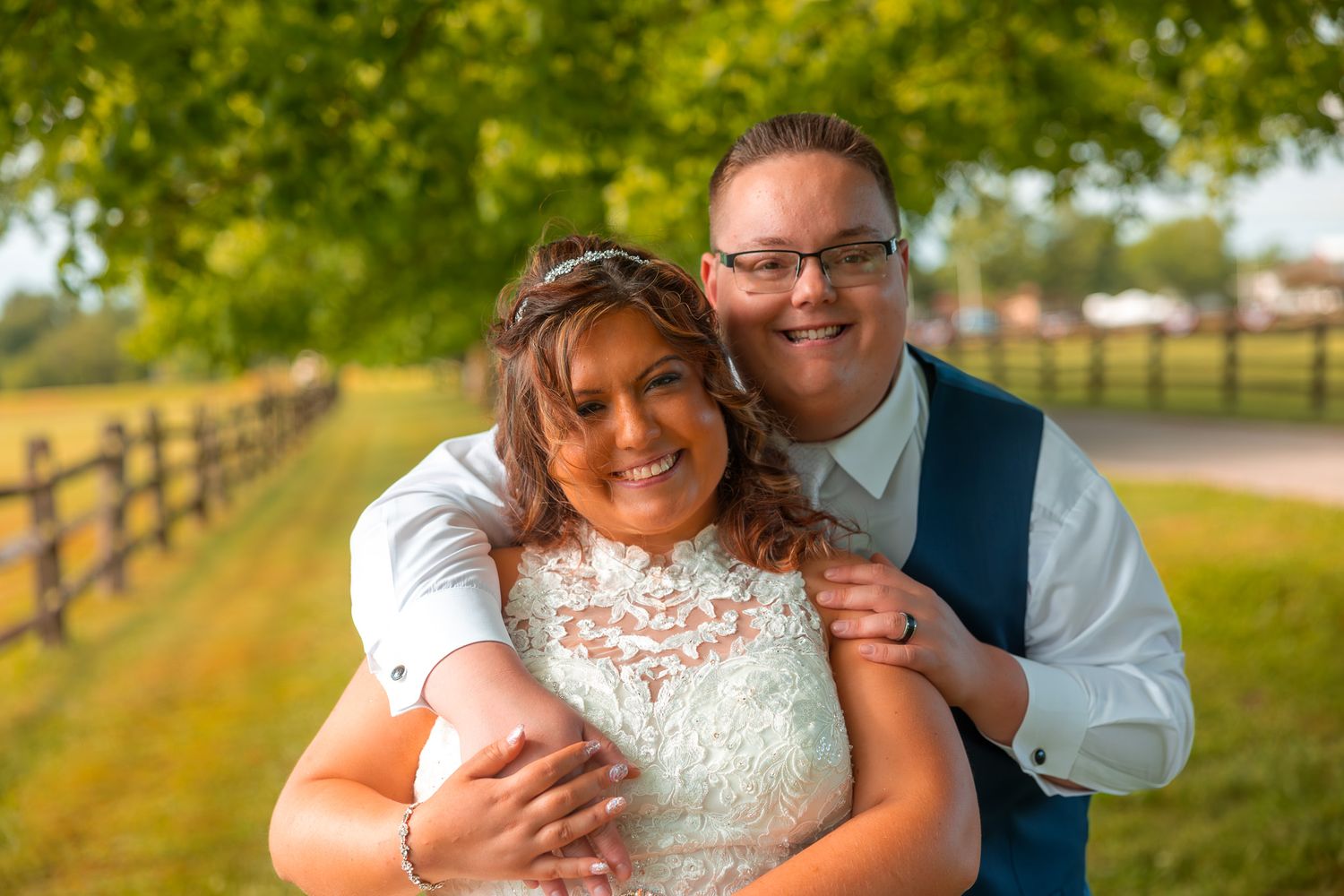 A happy couple poses together outdoors on their wedding day with a wooden fence and green trees in the background.