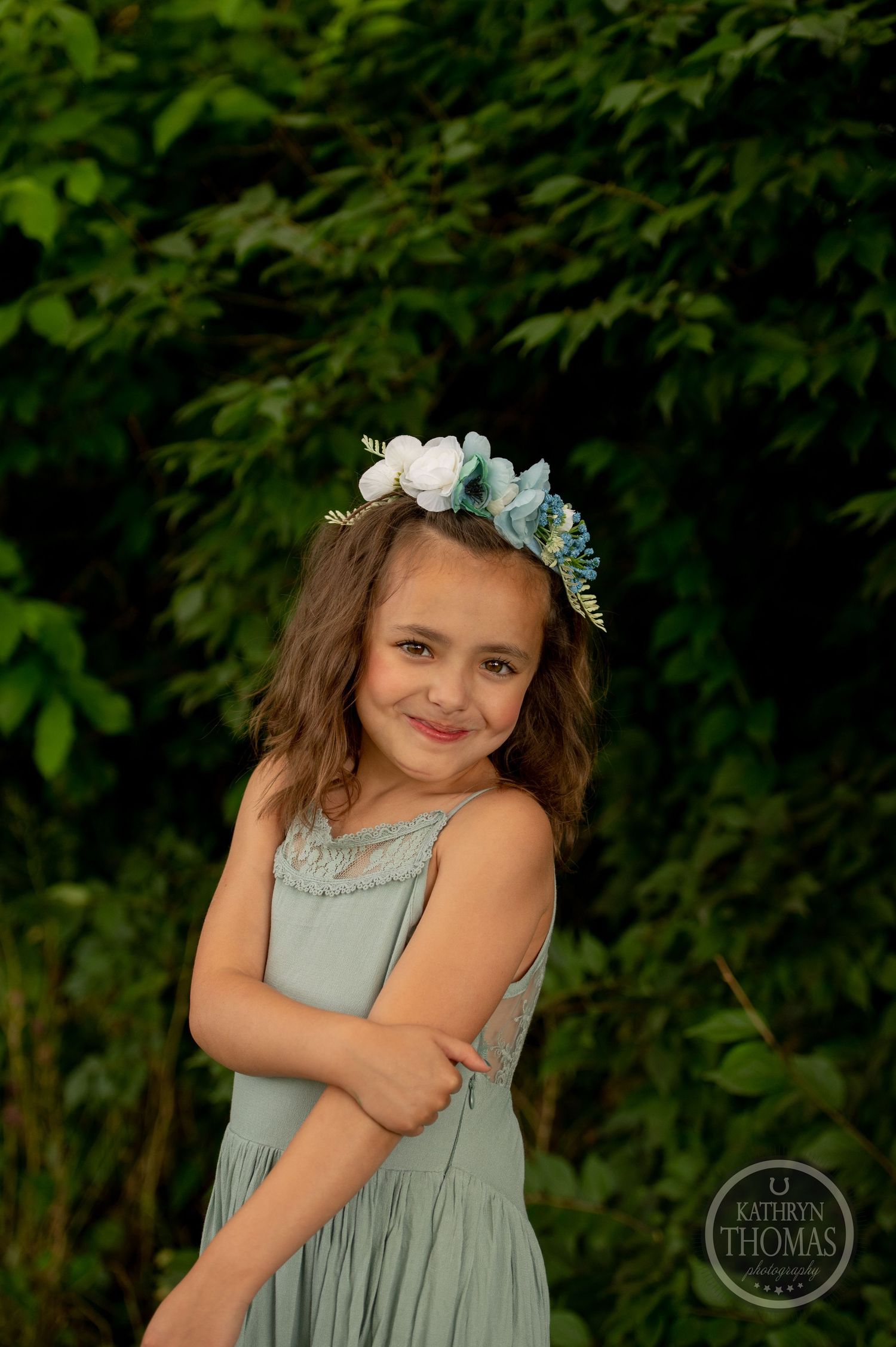 A whimsical portrait of a young child wearing a mint dress and floral crown against green foliage.