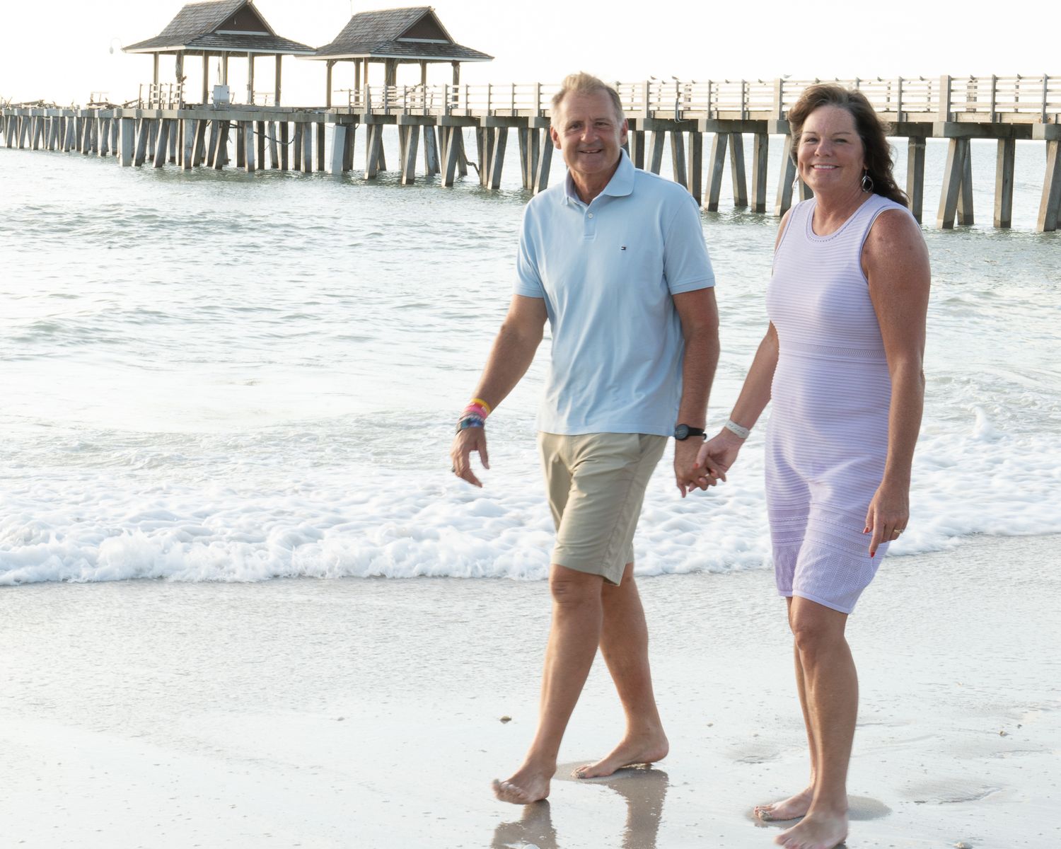A couple strolls hand in hand along a beach near a wooden pier at sunset.