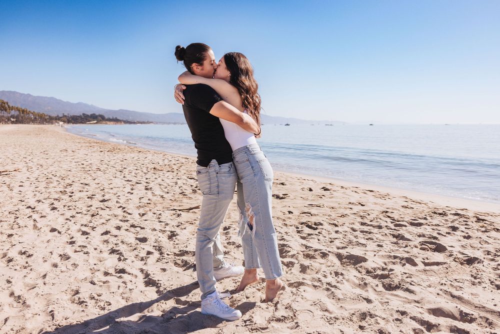 A couple in casual wear hugs and spins on an empty beach with mountains visible in the distance.