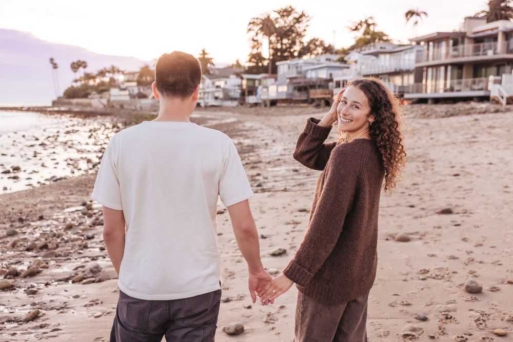 Two people walk together on a beach at sunset with oceanfront homes visible along the coastline.