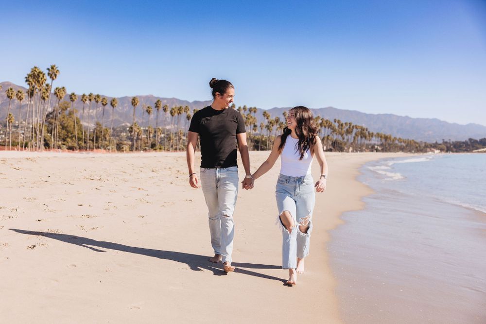 A couple walking together on a sandy beach with palm trees and mountains in the background during daytime.
