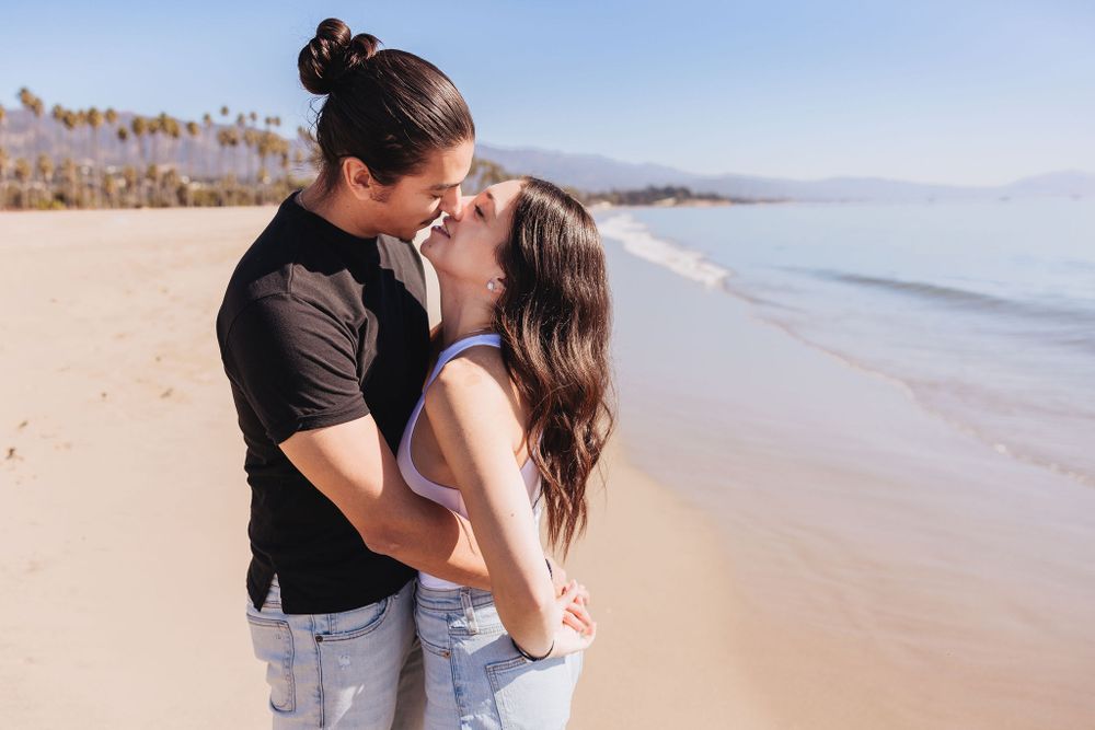 A couple embraces lovingly on a sunny beach with palm trees and ocean waves in the background.