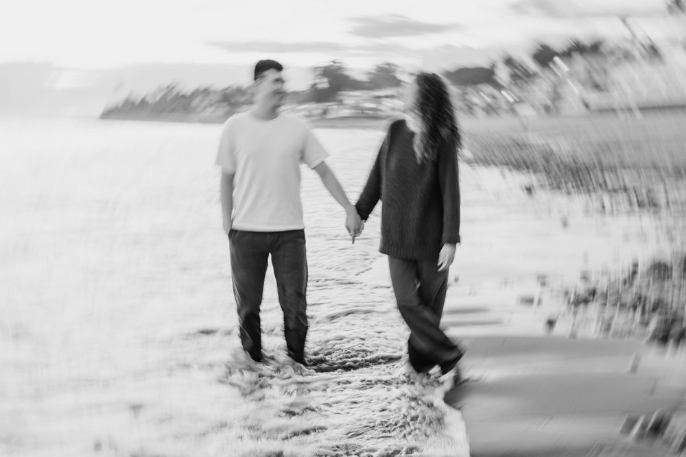 A black and white romantic photo of a couple holding hands while walking on a beach by the water.