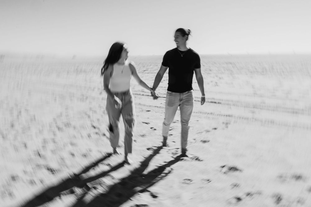 Black and white photograph of couple walking hand in hand on sandy beach casting long shadows on bright sand.
