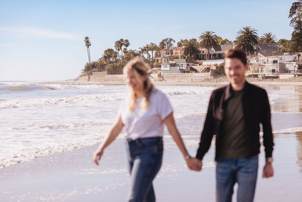 A couple walks along a sunny beach holding hands with coastal homes in the background.