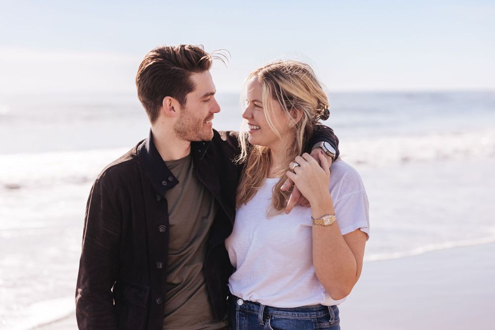 A joyful couple laughs together while embracing on a sunny beach, with the ocean visible in the background.