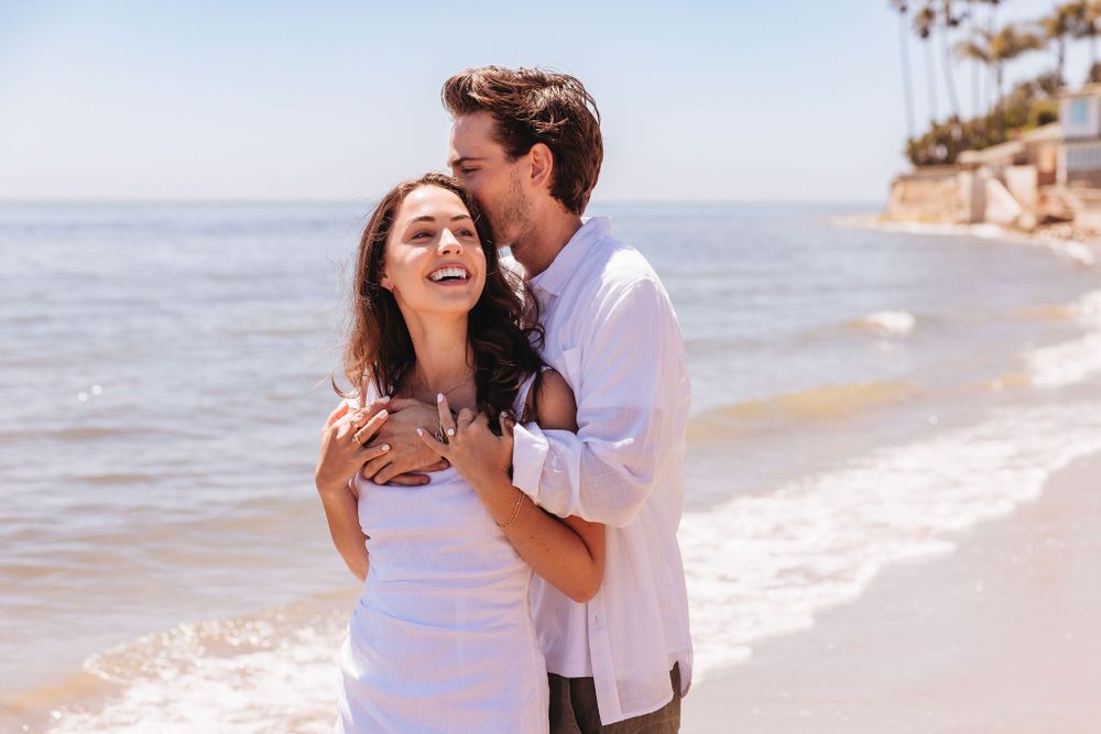 A joyful couple embracing and laughing together on a sunny beach with ocean waves in the background.