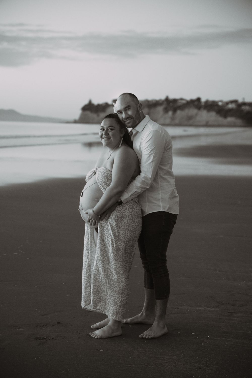 Couple poses together on beach for maternity photos at dusk with cliffs in background.